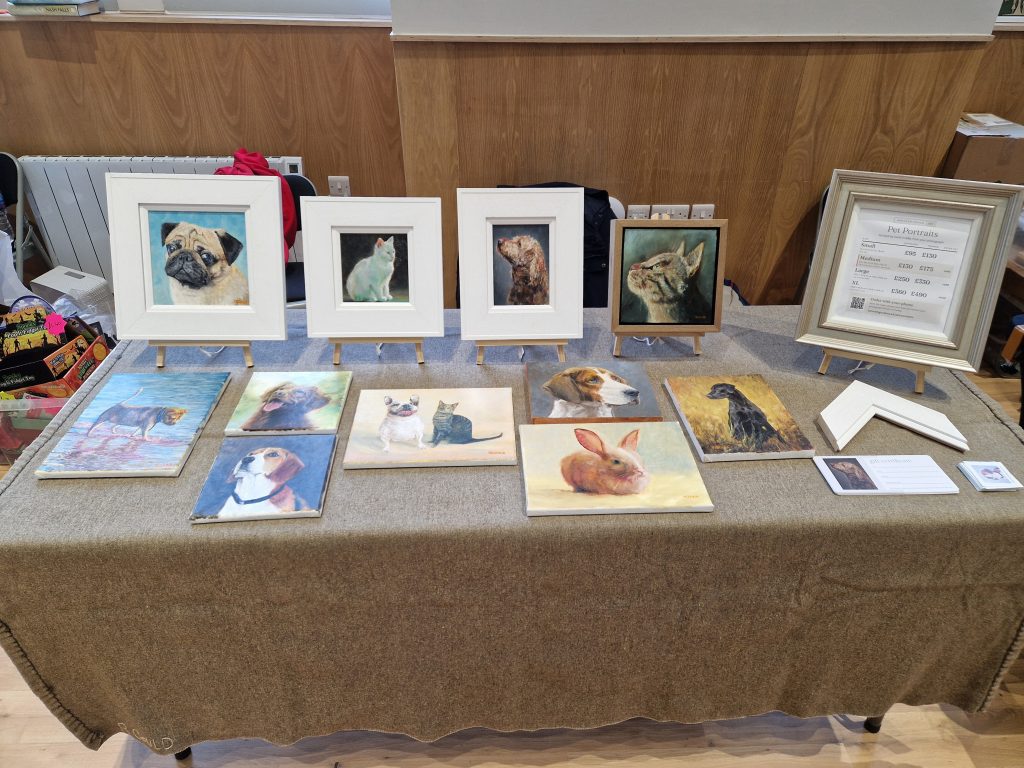 Kenneth Gould's table at Millport Town Hall: framed and unframed oil pet portraits on easels and laid on the table, with the pet portrait price board.